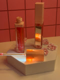 Diva's Love coral red lip gloss by Diva's Love Cosmetics, open with applicator displayed on white platform with mirror and pink crystals