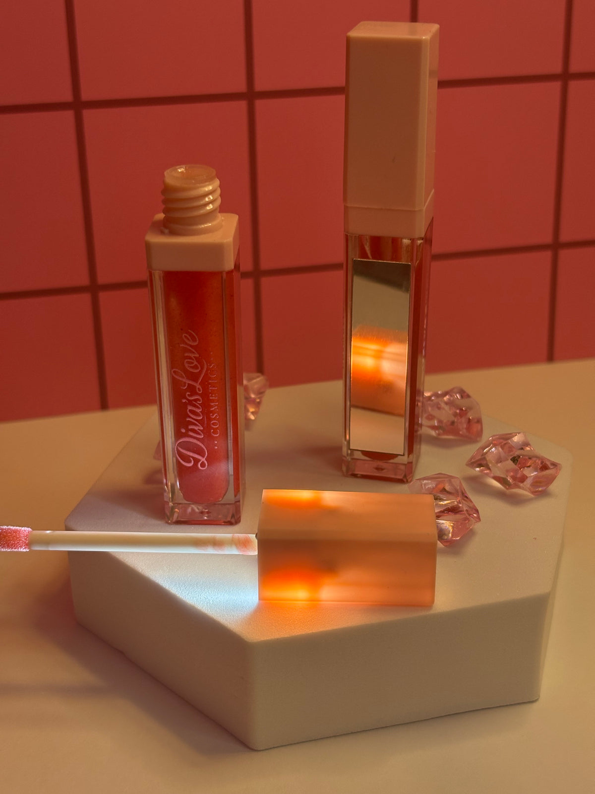 Diva's Love coral red lip gloss by Diva's Love Cosmetics, open with applicator displayed on white platform with mirror and pink crystals
